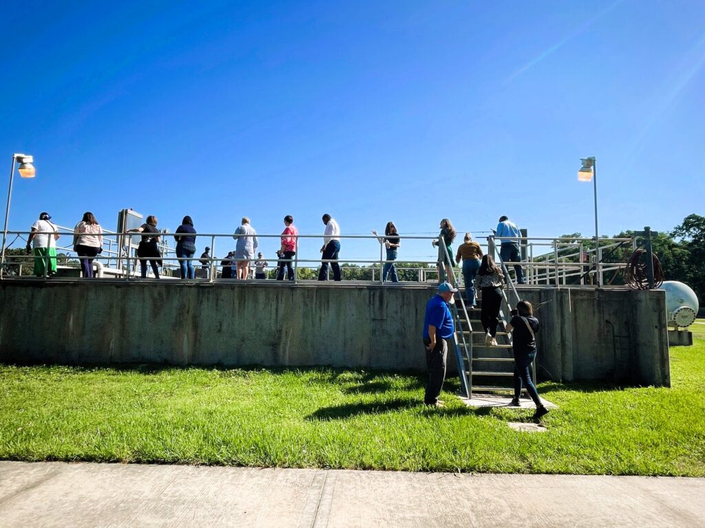 A group of people stands on a raised platform with railings at a water treatment plant, with stairs leading up from a grassy lawn under a clear blue sky.