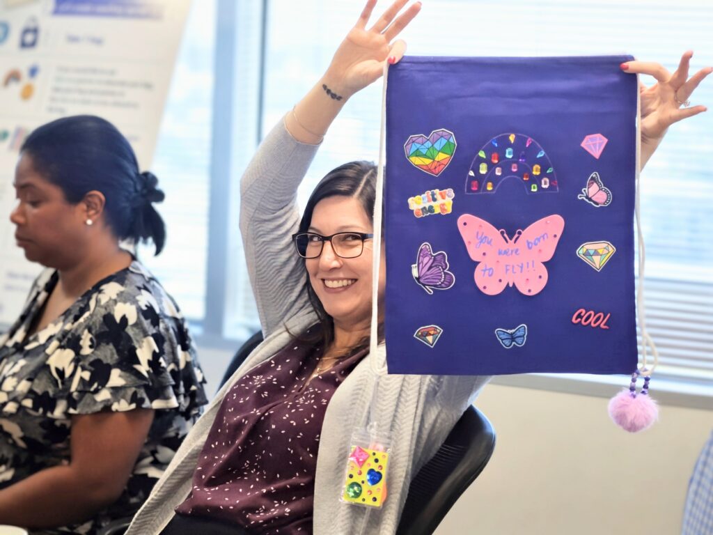 A woman in glasses holds up a purple decorated bag with butterfly stickers while seated in an office, with another woman working in the background.